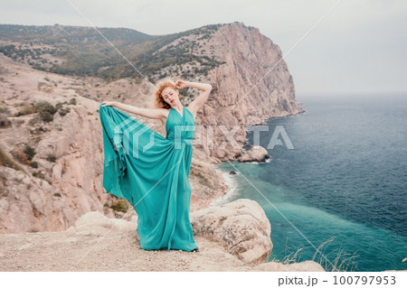 Redhead woman portrait. Curly redhead young caucasian woman with freckles looking at camera and smiling. Close up portrait cute woman in a mint long dress posing on a volcanic rock high above the sea Redhead woman portrait. Curly redhead young caucasian woman with freckles looking at camera and smiling. Close up portrait cute woman in a mint long dress posing on a volcanic rock high above the sea 100797953