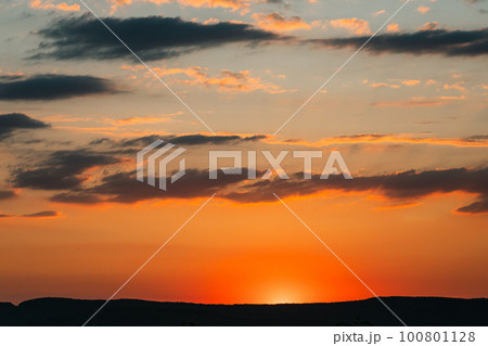 Sun hidden behind the silhouette of a mountain. Summer sunset in yellow and orange colours. The sun's rays from below beautifully illuminate the dark clouds. The beauty of nature. 100801128