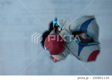 Close up of the face of a creepy clown looking through a hole. The face of a man wearing a creepy clown mask as he looks at his victim. Blue lighting. The clown madman. 100801130