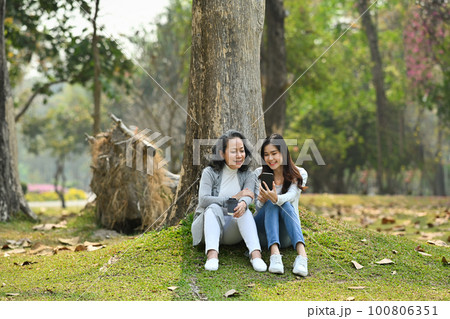 Photo of smiling senior mother and adult daughter using smart phone while sitting on green grass against calm nature park Photo of smiling senior mother and adult daughter using smart phone while sitting on green grass against calm nature park 100806351