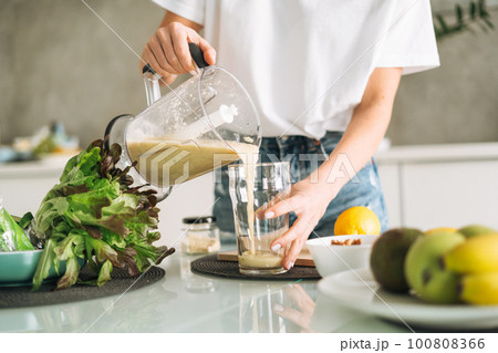 Young woman in white t-shirt and blue jeans cooking green smoothie healthy food in kitchen at home 100808366