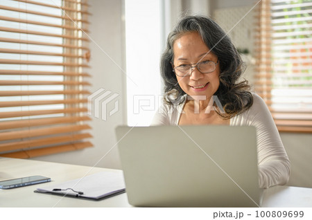 Smiling 60-year-old Asian businesswoman working on her business tasks on a laptop Smiling 60-year-old Asian businesswoman working on her business tasks on a laptop 100809699