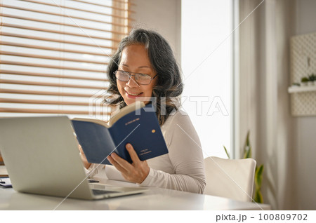 Relaxed and happy 60s Asian-aged woman reading a book at her workspace. 100809702