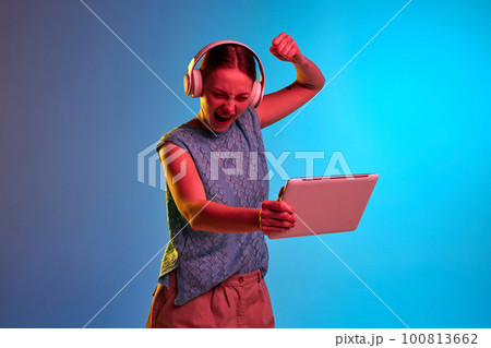 Excited young girl, holding tablet and hand up like symbol of win, screaming over blue background in neon light Excited young girl, holding tablet and hand up like symbol of win, screaming over blue background in neon light 100813662