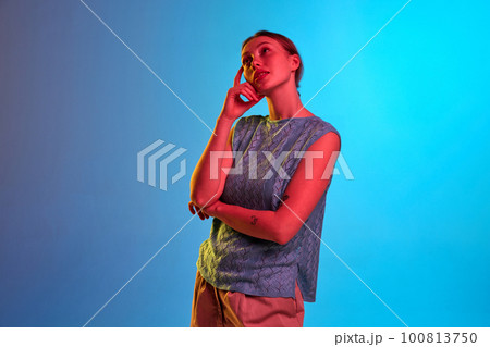 Shot of pensive girl with holded hands standing and thinking about something over blue background in neon light 100813750