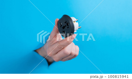 A woman is holding an outlet adapter. A woman's hand sticks out through a hole in a blue background. A woman is holding an outlet adapter. A woman's hand sticks out through a hole in a blue background. 100815941