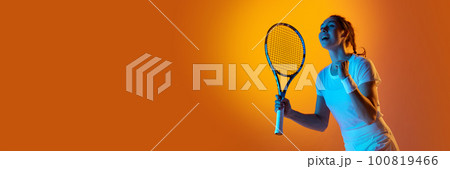 Happy winning emotions. Young female tennis player in uniform posing with racket against orange studio background in neon light Happy winning emotions. Young female tennis player in uniform posing with racket against orange studio background in neon light 100819466