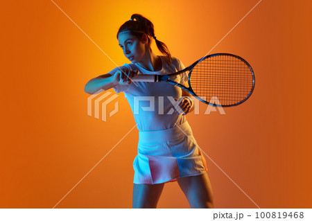 Portrait of young woman in white uniform, with tennis racket posing against orange studio background in neon light 100819468