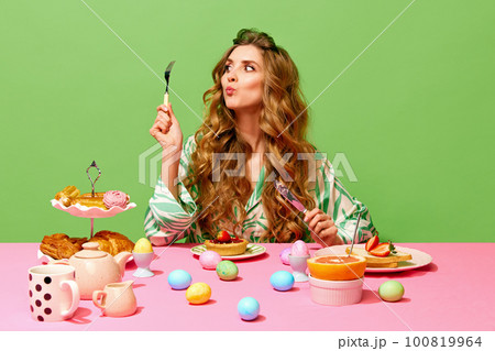 Food pop art of beautiful girl with long hair sitting at table holding cutlery and eating over green background. Colorful eggs on table 100819964