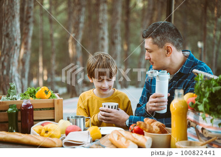 Father dad and school kid boy child having a picnic in the forest camping site with vegetables, juice, coffee, and croissants. Wooden crate with fresh organic veggies surrounded with bread baguettes. Father dad and school kid boy child having a picnic in the forest camping site with vegetables, juice, coffee, and croissants. Wooden crate with fresh organic veggies surrounded with bread baguettes. 100822441