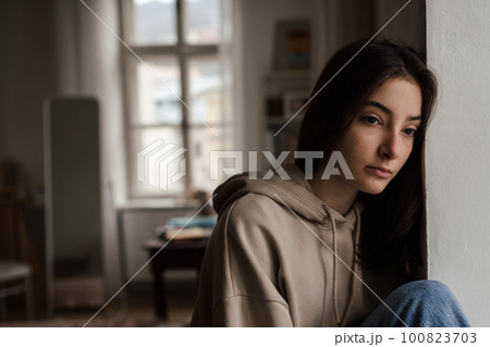 Unhappy teenage girl sitting in home and looking out of window. Unhappy teenage girl sitting in home and looking out of window. 100823703