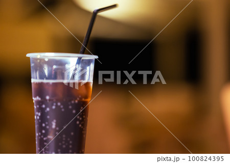 In Foreground In Focus Is Plastic Cup With Dark Cold Drink And Black Straw, In Background Out Of Focus Is Empty Beach Bar At Night. In Foreground In Focus Is Plastic Cup With Dark Cold Drink And Black Straw, In Background Out Of Focus Is Empty Beach Bar At Night. 100824395