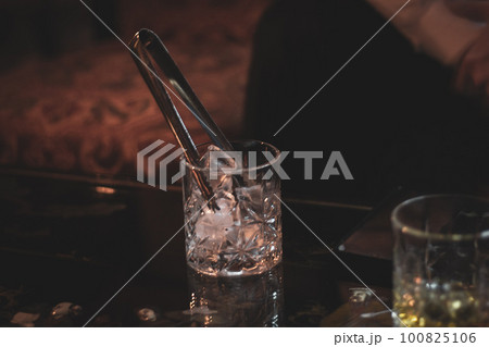 Whiskey glass with ice tongs. Bar counter concept. Brandy glass with ice cubes. Evening in bar. Alcoholic drinks. Cool alcohol drinks. 100825106