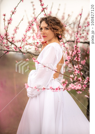 Woman peach blossom. Happy curly woman in white dress walking in the garden of blossoming peach trees in spring Woman peach blossom. Happy curly woman in white dress walking in the garden of blossoming peach trees in spring 100827120
