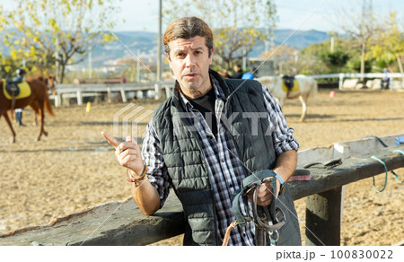 Portrait of skilled adult male stable worker holding horse halter or harness near wooden fence on ranch 100830022