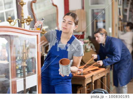 Young female furniture maker diligently working on vintage glass cabinet in workshop 100830195