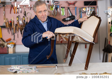 Elderly male furniture maker working on vintage chair in workshop Elderly male furniture maker working on vintage chair in workshop 100830201