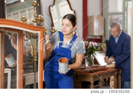Young female furniture workshop worker repainting vintage glass cabinet 100830280