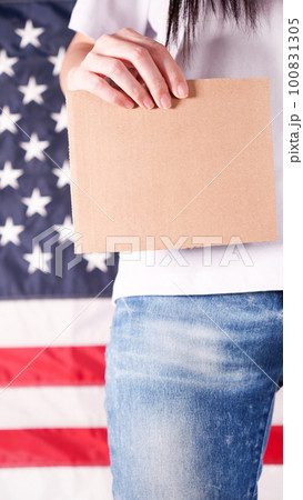 Young woman holds empty cardboard with Space for Text sign against American flag on background. Girl protesting anti-abortion laws. Feminist power. Womens rights freedom. Young woman holds empty cardboard with Space for Text sign against American flag on background. Girl protesting anti-abortion laws. Feminist power. Womens rights freedom. 100831305