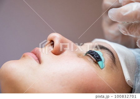 Close-up portrait of a woman on eyelash lamination procedure. The master applies tint to the eyelashes.  100832701
