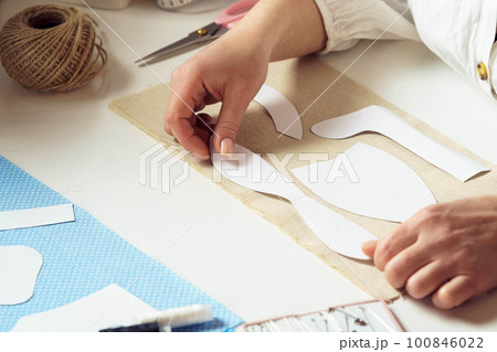 Unrecognizable closeup women, seamstresses holding and placing paper sew pattern, templates on textile sheet in workshop 100846022
