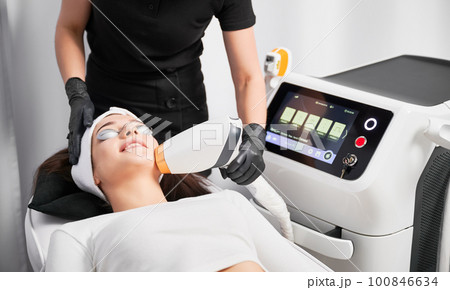 Female patient in safety glasses lying near cosmetology equipment and having face lifting procedure. Doctor cosmetologist using laser scanner device while performing facial treatment in beauty salon. Female patient in safety glasses lying near cosmetology equipment and having face lifting procedure. Doctor cosmetologist using laser scanner device while performing facial treatment in beauty salon. 100846634