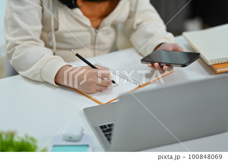 Cropped image of woman hand holding smart phone and writing records to daily planner  100848069
