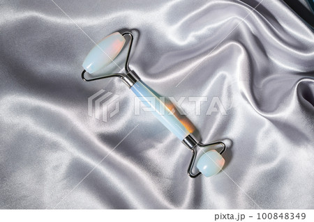 Blue jade face roller and Gua Sha scraping for beauty facial massage therapy on silver textured silk background. 100848349