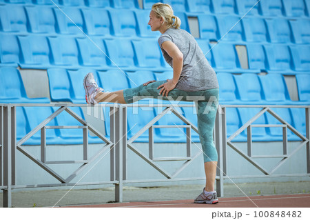 female athlete doing stretching exercise, tilt forward to leg raised on the fence, warm-up before athletics training female athlete doing stretching exercise, tilt forward to leg raised on the fence, warm-up before athletics training 100848482
