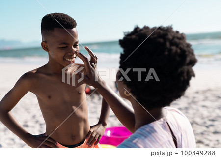 African american mother applying sunscreen on son's nose at beach against clear sky, copy space 100849288