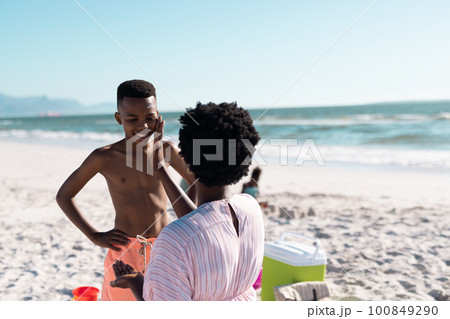 African american woman applying sunscreen on son's face at beach against clear sky, copy space 100849290