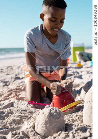 Playful african american boy building a sandcastle with pail on beach against clear sky, copy space 100849298