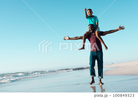 Cheerful african american man with arms outstretched carrying son on shoulders at seashore under sky Cheerful african american man with arms outstretched carrying son on shoulders at seashore under sky 100849323