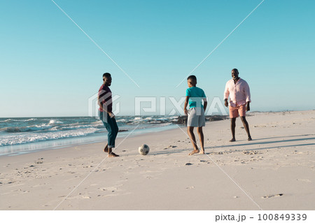 African american boy playing soccer with father and grandfather at beach against clear sky 100849339