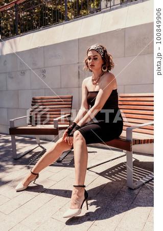 Portrait of a woman on the street. An attractive woman in a black dress is sitting on a bench outside. Portrait of a woman on the street. An attractive woman in a black dress is sitting on a bench outside. 100849906