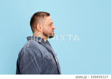 Profile view of one nice-looking man wearing jeans jacket standing with calm facial expression over blue background. Side view 100856127