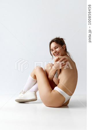 Portrait of positive, smiling, slim young girl covering breast with hand, posing in underwear against grey studio background. 100856456