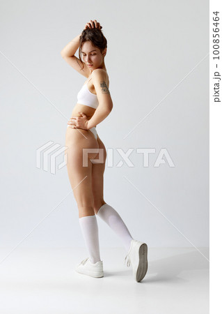 Full-length side view image young girl with slim, fit body posing in underwear, high socks and sneakers against grey studio background. Full-length side view image young girl with slim, fit body posing in underwear, high socks and sneakers against grey studio background. 100856464