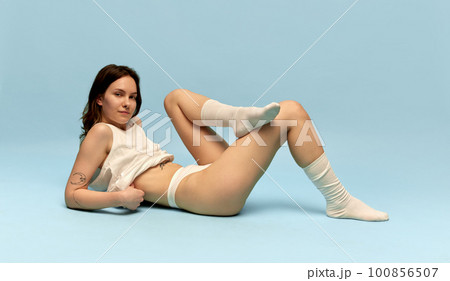 Portrait of beautiful young girl with slim body, in comfortable homewear and socks posing against blue studio background. Coziness Portrait of beautiful young girl with slim body, in comfortable homewear and socks posing against blue studio background. Coziness 100856507