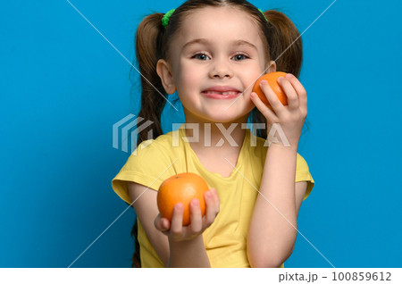 A small and smiling girl is holding two tangerines on a blue background. 100859612