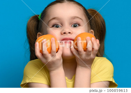 A small and smiling girl is holding two tangerines on a blue background. A small and smiling girl is holding two tangerines on a blue background. 100859613