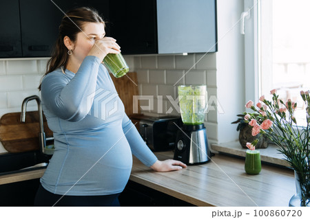 Green Smoothies Recipes For Pregnancy and Postpartum, Prenatal Nutrition. Pregnant woman preparing green vitamin smoothie with a blender and drinking in the kitchen. 100860720