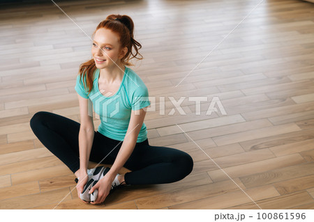 High-angle view of smiling young yogini woman practicing yoga, sitting in butterfly konasana pose, stretching muscles on floor at home. Pretty female sitting on floor in lotus butterfly yoga pose High-angle view of smiling young yogini woman practicing yoga, sitting in butterfly konasana pose, stretching muscles on floor at home. Pretty female sitting on floor in lotus butterfly yoga pose 100861596