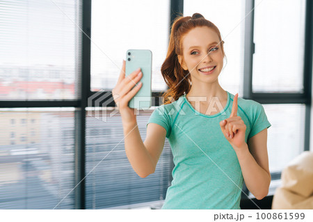 Portrait of happy redhead young woman in casual clothes talking on video call holding smartphone in hand shooting selfie on front camera and showing index finger up, standing on background of window Portrait of happy redhead young woman in casual clothes talking on video call holding smartphone in hand shooting selfie on front camera and showing index finger up, standing on background of window 100861599