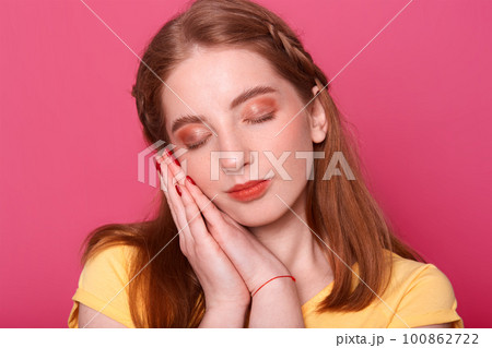 Indoor studio shot of romantic pretty young lady with closed eyes and hands near face, going to have nap, looks sweet. Attractive model poses isolated over bright pink background. Youth concept. 100862722