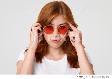 Concentrated young European female with serious expression, keeps hands on temples, tries to focus on something, wears red sunglasses and white casual t shirt, isolated over studio background Concentrated young European female with serious expression, keeps hands on temples, tries to focus on something, wears red sunglasses and white casual t shirt, isolated over studio background 100863019