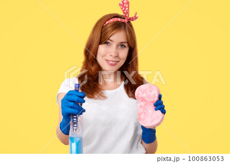 People and housekeeping concept. Adorable smiling female housewife in casual clothes, does house duties, cleans in room, holds bottle with detergent and sponge, stands over yellow background People and housekeeping concept. Adorable smiling female housewife in casual clothes, does house duties, cleans in room, holds bottle with detergent and sponge, stands over yellow background 100863053
