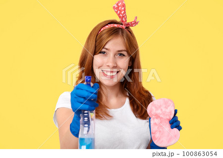 Lovely young housemaid in headband, rubber protective gloves, uses washing spray and sponge for cleaning mirror, smiles positively, isolated over yellow background, rejoices finishing housework 100863054