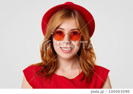 Close up shot of pretty Caucasian young female with gentle smile, looks happily at camera, wears red shades and hat, rejoices recieving compliment, isolated on white background. Fashion concept Close up shot of pretty Caucasian young female with gentle smile, looks happily at camera, wears red shades and hat, rejoices recieving compliment, isolated on white background. Fashion concept 100863096