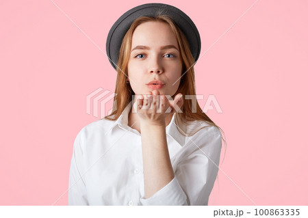 Portrait of beautiful female blows air kiss at camera, expresses her love, wears hat and shirt, flirts with boyfriend during date, stands against pink studio background. This kiss is for you! 100863335